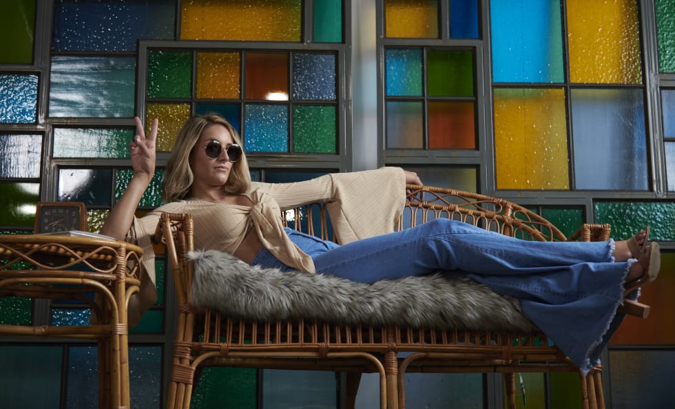Beautiful young woman in attired from 1960Õs (flared shirt and bell bottom jeans) sits on chair in front of stained glass holding peace sign